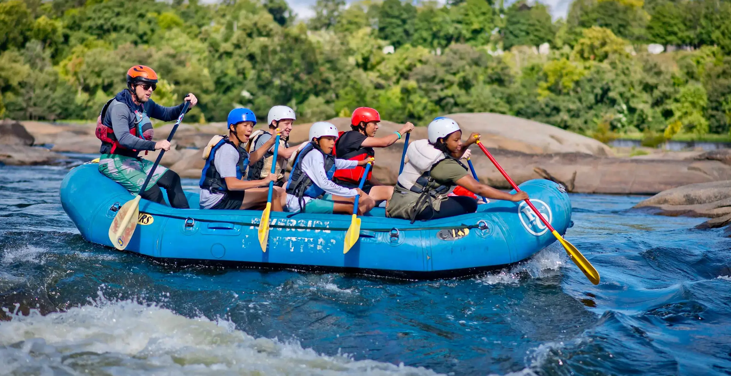 Students wearing helmets and life jackets smiling while paddling together in a raft on the river.